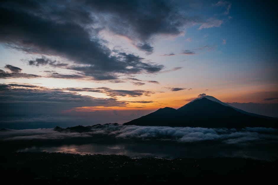 Serene view of Mount Batur silhouetted against a colorful sunset sky, surrounded by clouds and reflecting water.